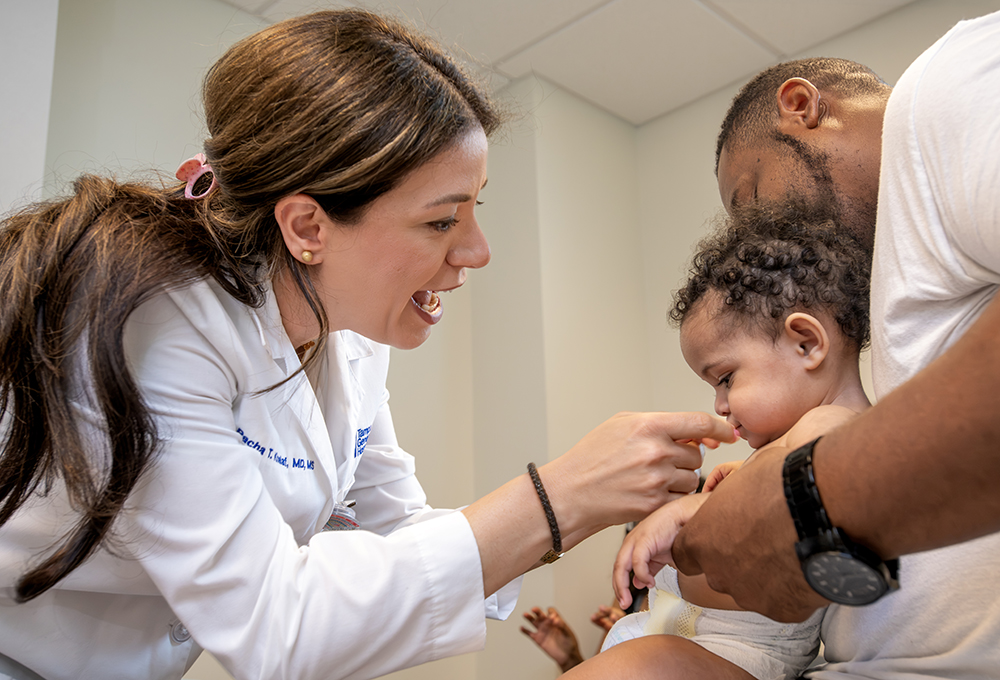 Dr. Racha Khalaf, wearing a white coat, smiles and reaches toward a toddler held by her father in a clinic exam room.