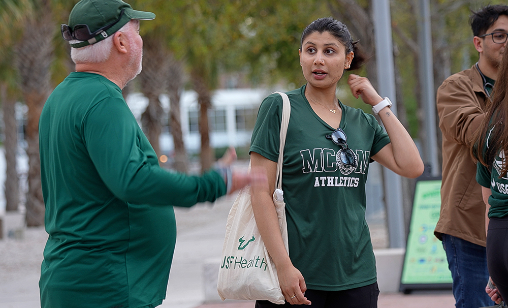 Two people stand outdoors in conversation. On the left, an older man wearing a green long-sleeve shirt and green cap gestures while speaking. On the right, a young woman wearing a green MCOM Athletics shirt and carrying a USF Health tote bag listens and responds.