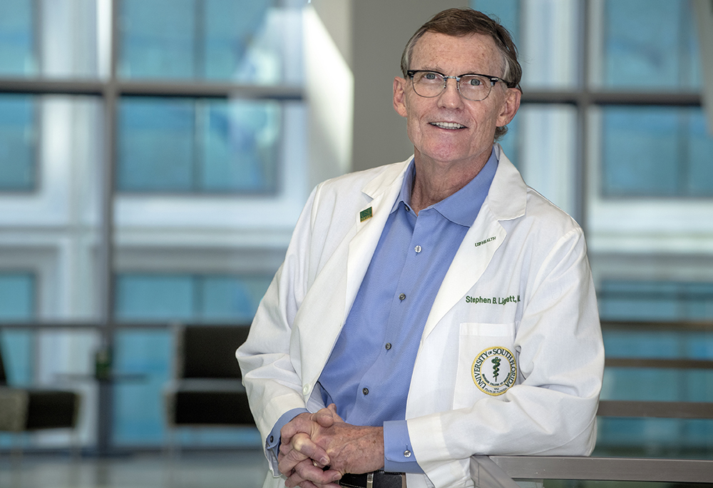 Dr. Stephen Liggett wears glasses and a white medical coat over a blue shirt, standing in front of modern glass windows. He has his hands clasped together and is smiling at the camera in a professional portrait taken in the MDD building in downtown Tampa.