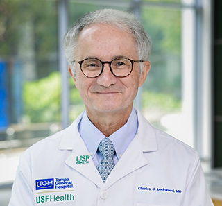 An older man in a white medical coat smiles at the camera wearing brown eyeglasses in a mult-window lobby