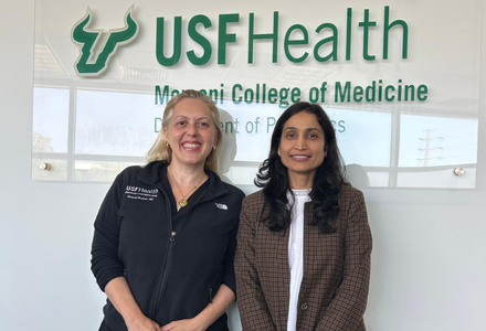 Two women stand posing in front of a USF Health sign