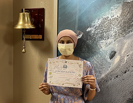 A young woman wearing a pink head covering and surgical mask stands next to a brass bell mounted on a wall. She holds a congratulations certificate signed by her care team from the Moffitt Infusion Center recognizing her completion of cancer treatment.