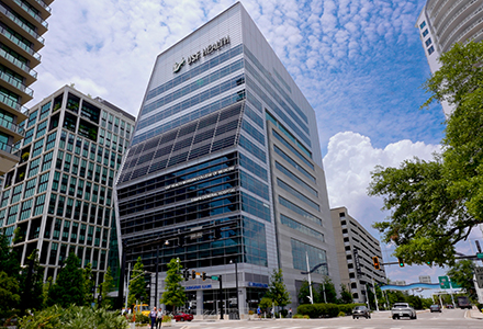 The exterior of the USF Health Morsani College of Medicine building in downtown Tampa, a modern glass tower bearing the USF Health name, viewed from a street-level intersection on a partly cloudy day.