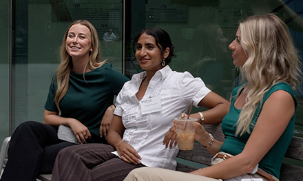 Three women sit together outdoors, smiling and talking in a relaxed setting against a glass building exterior.