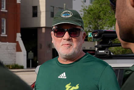 An older man with a white beard wearing a green USF t-shirt, green cap and sunglasses stands outdoors, speaking with people partially visible on either side of him.