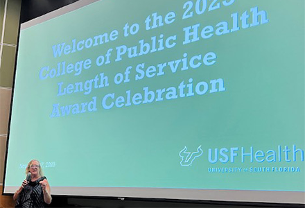 Dean Donna Petersen stands in front of a powerpoint presentation that reads "Welcome to the 2023 College of Public Health Length of Service Celebration", holding a microphone
