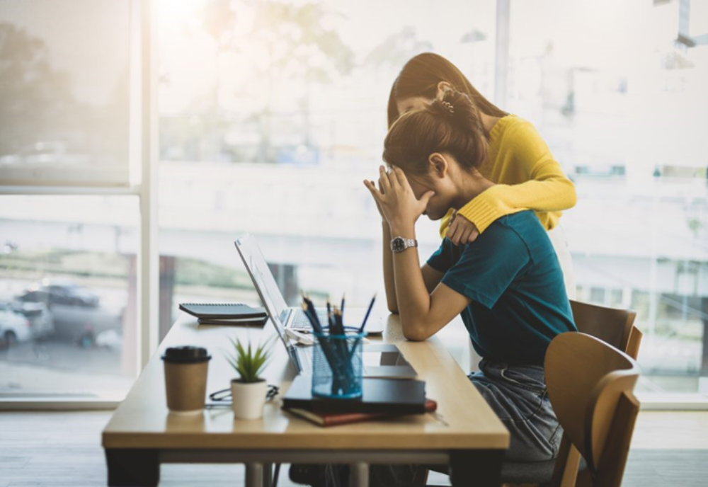 A co-worker comforts a colleague in distress at her desk.