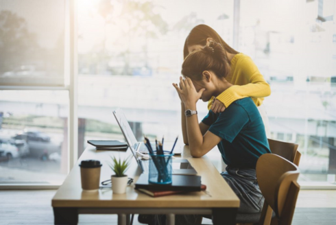 A co-worker comforts a colleague in distress at her desk.