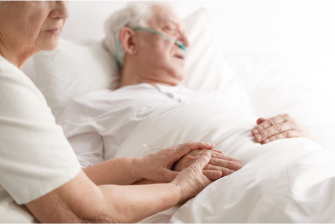 Older man lying in hospital bed and older woman holds his hand.