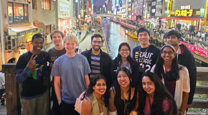 A group of Honors students pose at night on a bridge overlooking a canal and brightly-lit buildings.