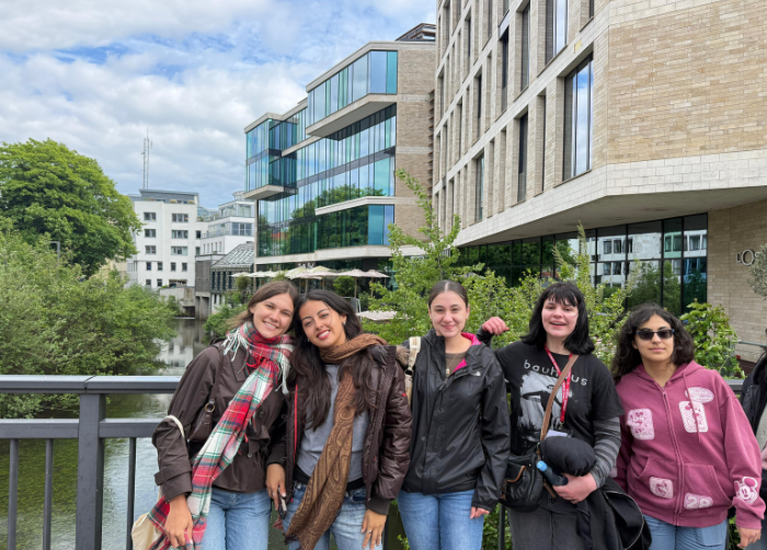 Group of five female students leaning on the bridge railing with buildings and a river behind them.