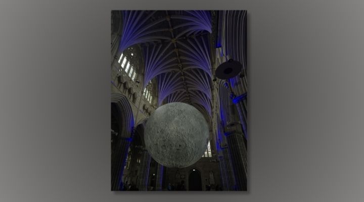 A photo taken inside a dark, gothic cathedral in Exeter with an art installation of a giant, hanging moon above the pews
