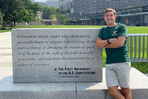 Samuel Rechek wearing commencement regalia sit in front of a University of South Florida sign