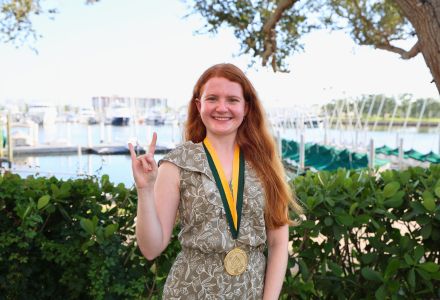 Elizabeth Delp with the "Go Bulls" handsign an Honors medallion on the USF St. Petersburg campus with water and boats in the background.