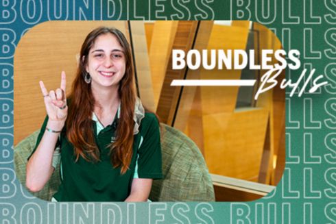 May Birch, posing with the Go Bulls hand sign while sitting in a chair overlooking the Honors atrium.