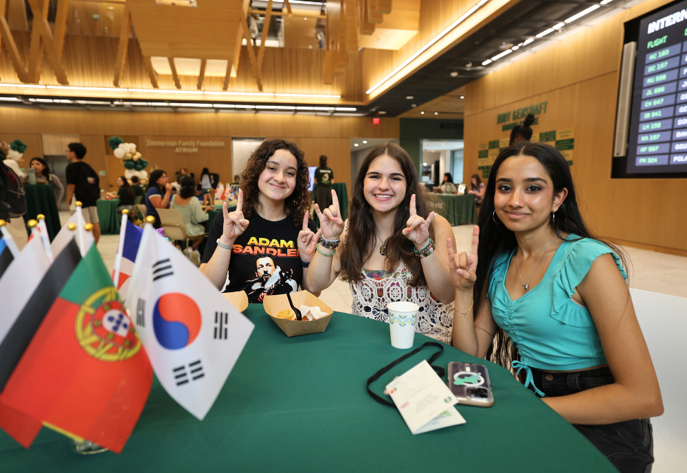 Three Honors students show their bull pride in the Honors atrium at a round table with a green tablecloth and a multi-cultural flag centerpiece.