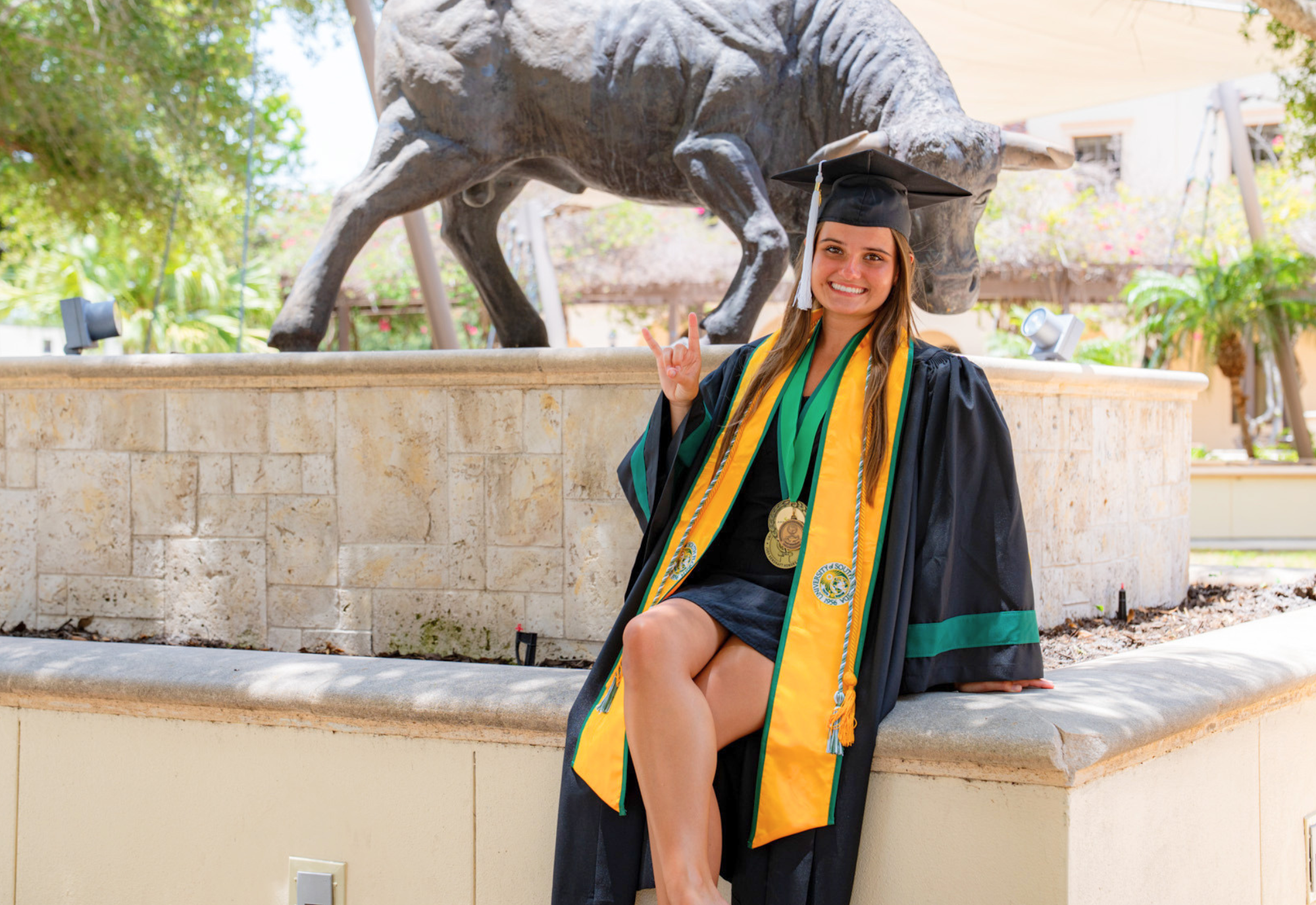 Britlee Yant in USF cap and gown holding up the Go Bulls hand sign with a bull statue in the background