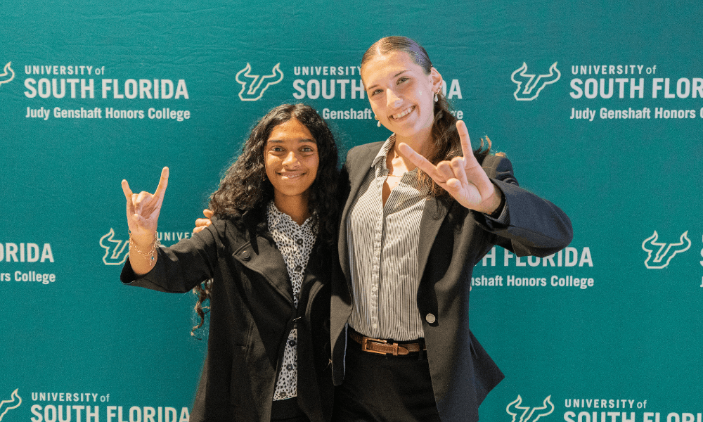 Katelyn Carlsen and Sarah Ismail hold up "Go Bulls" hand sign in front of a green USF Honors backdrop