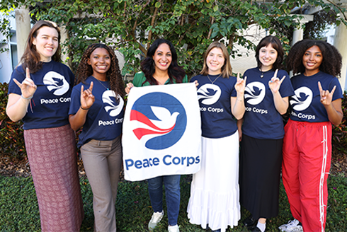 Six people stand with Peace Corps shirts on and the center person holding up a banner with the Peace Corps logo and text.