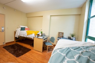 Bed on left with striped comforter, desk in front. Another bed on the other corner of the room.