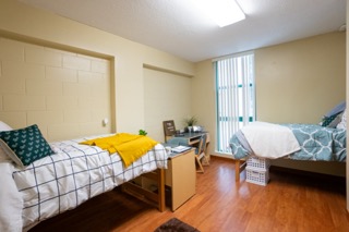 Different angle with bed on left with striped comforter, two desks by the window. Another bed on the other corner of the room.