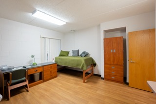 Desk on left and bed with green comforter on left. A large wardrobe is next to the bed.