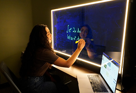 girl sits at lightboard while writing a math equation