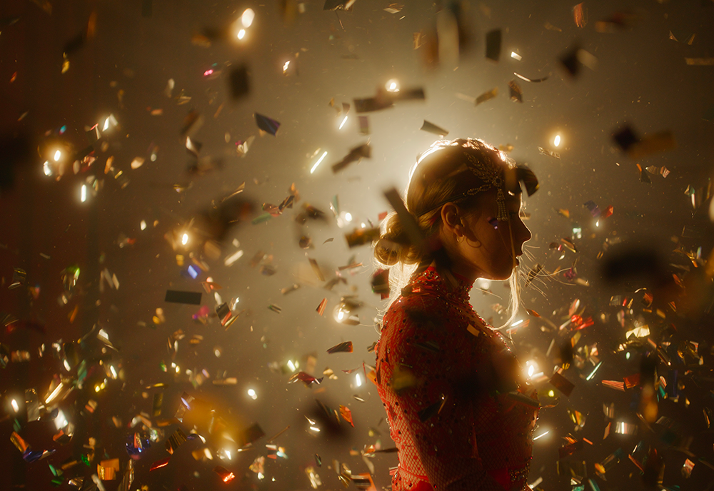 a woman circus performer stands with a bright spotlight behind her head and gold confetti all around her