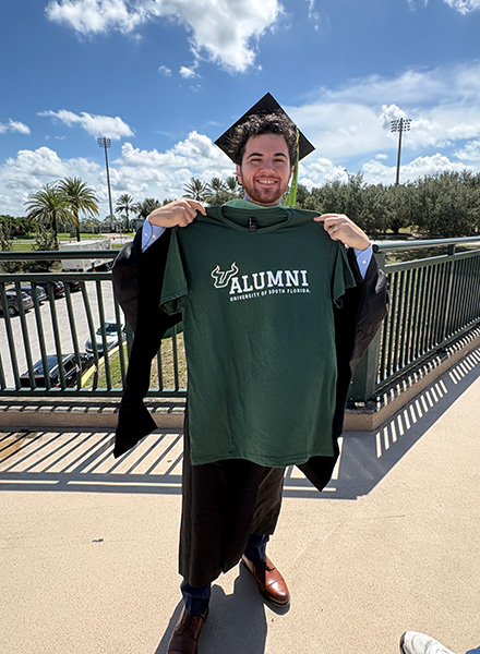 standing at graduation holding up alumni t-shirt