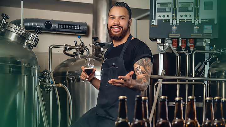 A male USF student holding a beer and standing in front of brewing equipment at the USF St. Petersburg campus
