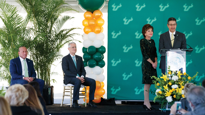 Judy Genshaft speaking at a podium while President Currall sits next to her