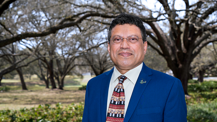 Provost Prasant Mohapatra, wearing a dark blue suit with a USF bull horn pin displayed, pictured outdoors in front of live oak trees at the Patel Center on USF’s Tampa campus.