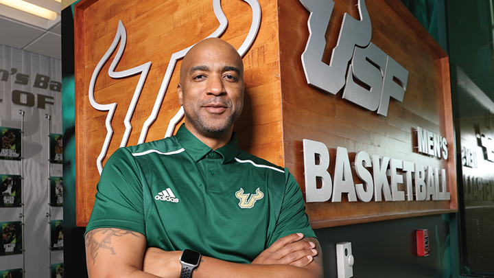 USF’s 11th men’s basketball head coach Amir Abdur-Hahim stands in front of Men’s Basketball signage in a USF Athletics building.