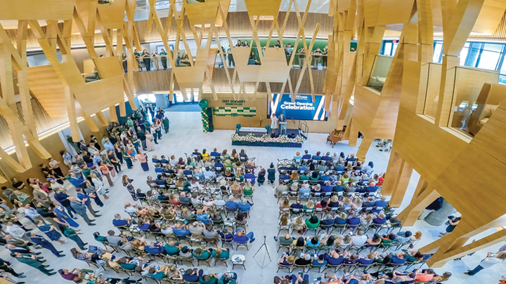 A overhead view of a large seated crowd in the beautiful atrium of the new Honors College building. President Emerita and husband Steven Greenbaum speak at a podium at the front of the room, with a Grand Opening digital screen behind them.