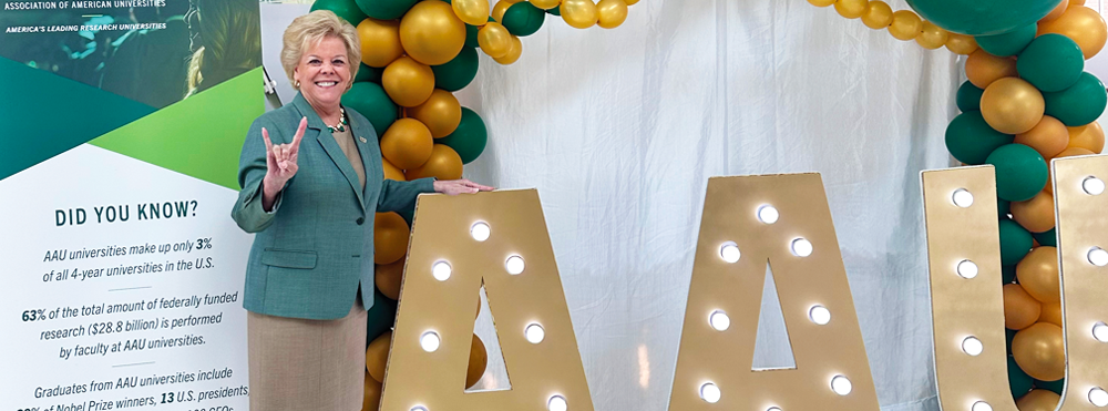 USF President Rhea Law smiles and makes the Bull U sign while standing by a display that includes 3 ft. tall gold AAU letters, with gold and green balloons.