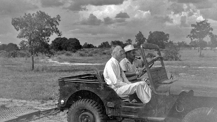 A historic black and white photo of USF’s first president John Allen riding with another man in an open jeep, surveying the undeveloped site for USF’s Tampa campus, scrub grass, sand and trees on a dirt road.