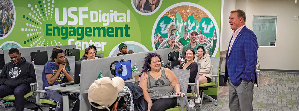 Several students chat with USF Foundation CEO Jay Stroman in the USF Digital Engagement call center on the Tampa campus. A large apple-green mural with photos of USF students grace the walls. 