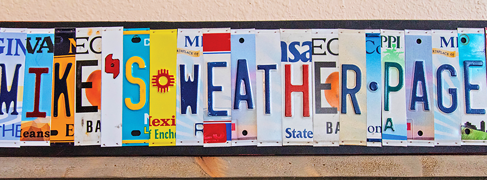 A colorful metal sign made from varied state license plates, with the text Mike’s Weather Page, is displayed on a wooden shelf. 