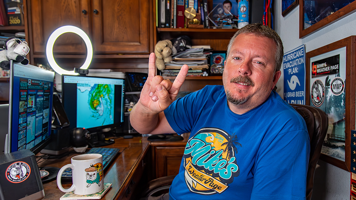 Mike Boylan, in royal blue T-shirt emblazoned with “Mike’s Weather Page, seated at his computer in an office surrounded by hurricane-chasing memorabilia.