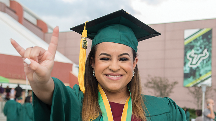 A smiling female USF graduate shows the Bull horn U gesture, in USF regalia on graduation day, in front of the Yuengling Center.