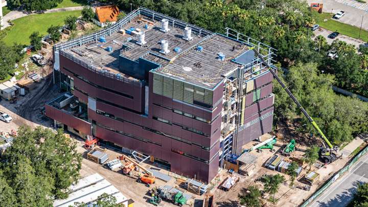 An aerial view of the mauve Honors College building under construction, showing a tall crane reaching the top of the five story building, surrounded by tall green trees. 