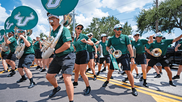 Bright midday sunlight shines on members for the Herd of Thunder brass section dressed in black shorts with USF polos and baseball caps.