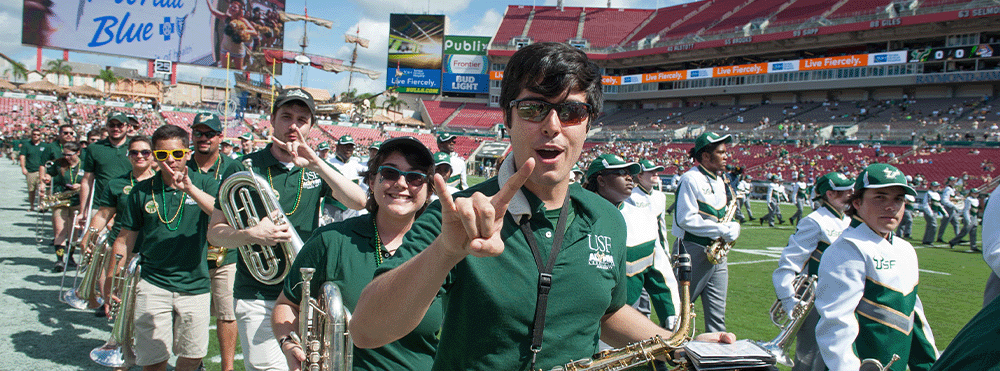 Current and alumni members of the Herd of Thunder line up on the sideline at Raymond James stadium for a performance during the Homecoming football game. An enthusiastic alumnus throws up the Bull horns hand gesture and looks right at the camera.