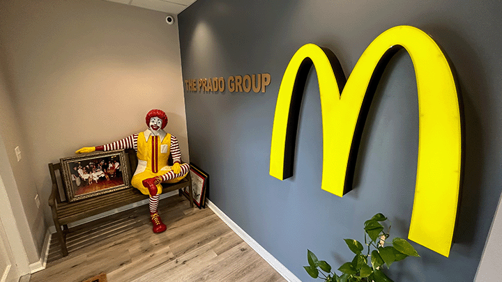 McDonald’s golden arches logo is on a gray wall next to gold lettering that reads “The Prado Group.” A park bench sits perpendicular to the wall with a seated life-sized statue of Ronald McDonald. His arm is draped around a framed family photo.