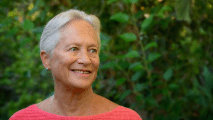 A portrait of Rebecca Penneys. Her gray hair is combed back from her face and she’s wearing a coral top. She stands in front of a manicured green hedge.