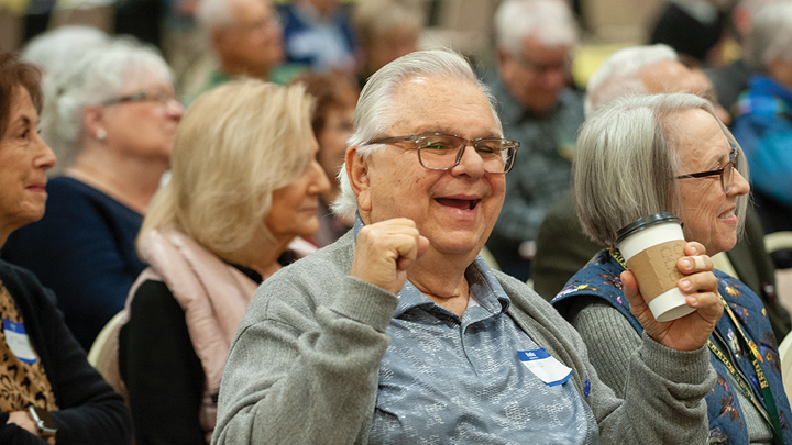 A large crowd of seated senior citizens listen to a presentation. An older man in a gray sweater and polo shirt smiles and holds a paper coffee cup in the foreground. 