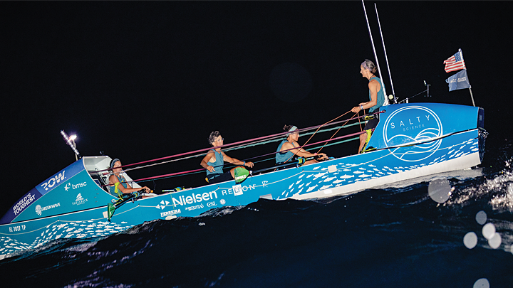A blue and white, large row boat tilts at a 20-degree angle on black waves at night. Three teammates face right, and the fourth faces her team, looking left. Two teammates are rowing, and a small American flag and World’s Toughest Row flag flutter in the breeze.