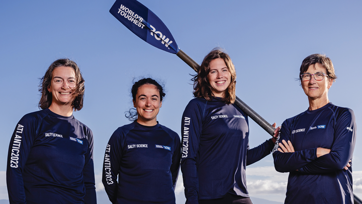 The four teammates, ages 20s to 40s, pose in dark blue team outfits emblazoned with the team name - Salty Science – and Atlantic 2023. The woman third from left holds a paddle over her shoulder, and the ocean and blue sky are behind them.