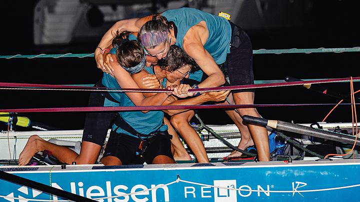 The four teammates embrace each other in a tight group hug, standing in their boat, at night. They are deeply tanned and wearing aqua blue tops and black shorts.