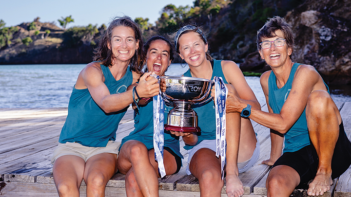 Four smiling and laughing women wearing aqua blue sleeveless shirts and shorts sit atop a wooden dock. Their bare feet are in the water, and they hold a large, silver trophy cup together.